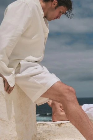 Man wearing a white linen shirt and shorts sitting on a low white wall against an ocean and sky background.