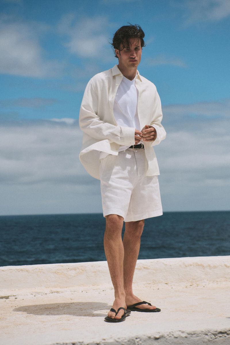 Man standing by the ocean in an open off-white shirt, white t-shirt, white shorts, and black leather sandals.