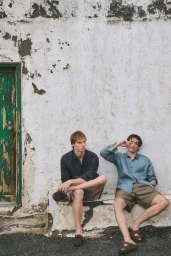 Two male models in linen button-down shirts and shorts seated against a textured white wall beside a green wooden door frame.
