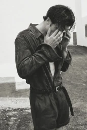 Monochrome photograph of a man in a suede button-down shirt and belted shorts covering his face outdoors.