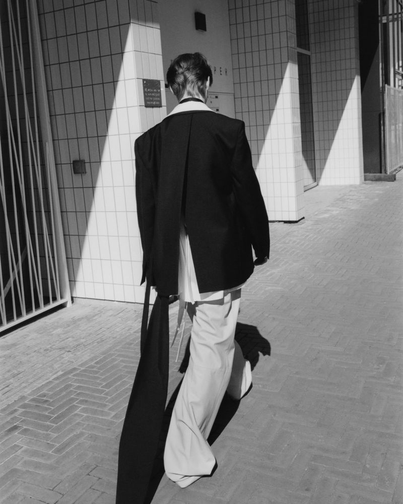 Person walking in black blazer and white pants on a brick walkway next to a tiled building with sunlight creating geometric shadows.