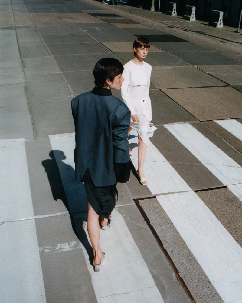 Two women in fashionable attire walk on a crosswalk in an urban environment on a sunny day, captured at eye-level with ample sunlight.