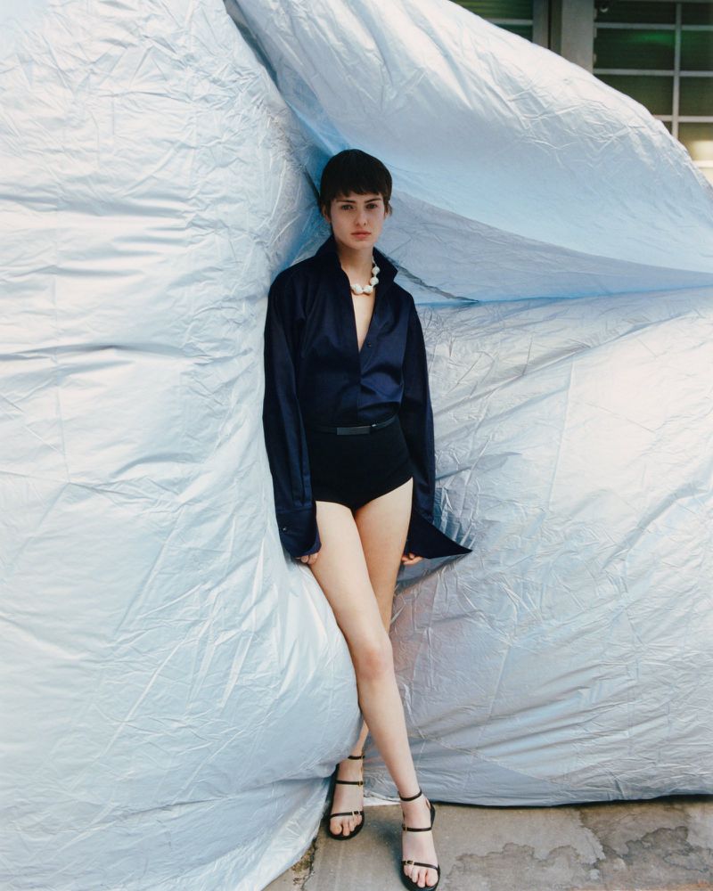 Young adult woman poses in a navy blouse and black bottoms with black strappy heels in front of a blue tarp outdoors.