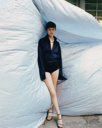 Young adult woman poses in a navy blouse and black bottoms with black strappy heels in front of a blue tarp outdoors.