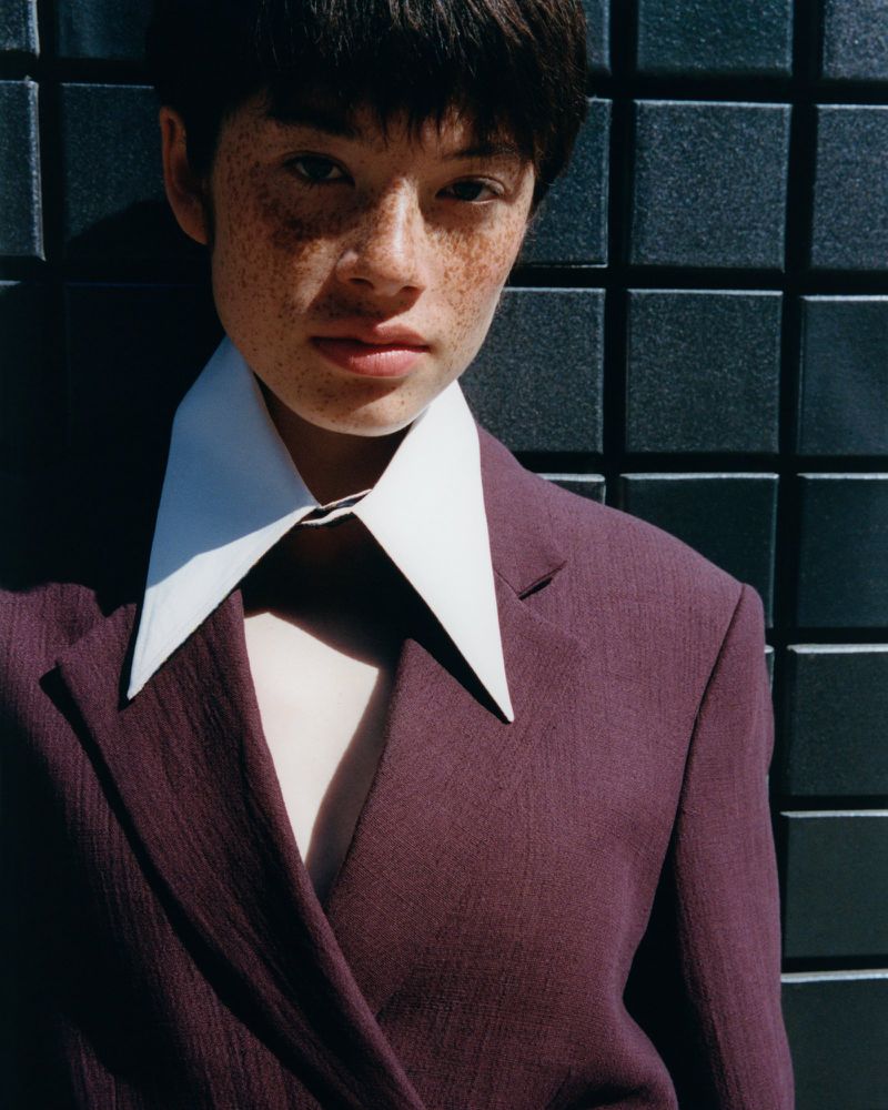 Close-up portrait of a young person with freckles, wearing a purple blazer with a white collar, against a dark tiled background.