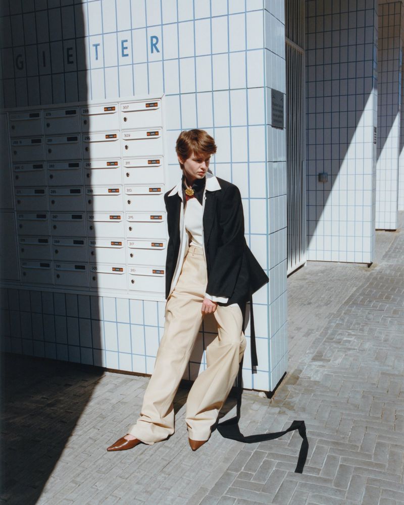 A woman in a black blazer and tan pants poses outside a tiled building, casting a long shadow on the stone-tiled ground during the day.