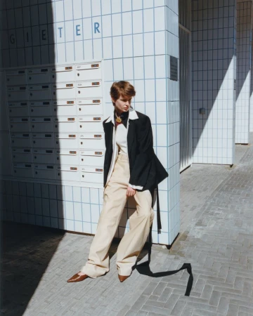 A woman in a black blazer and tan pants poses outside a tiled building, casting a long shadow on the stone-tiled ground during the day.