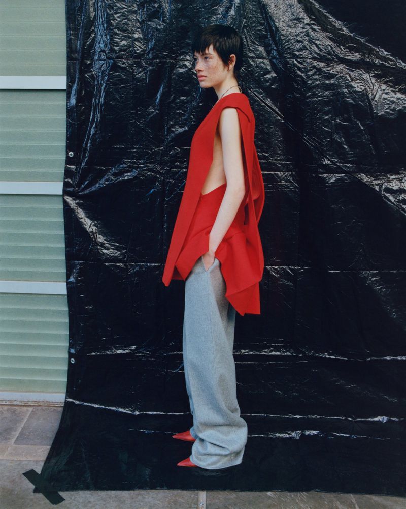 A side-view studio shot of a female model in a red draped top and loose grey pants, posing in front of a textured black backdrop.