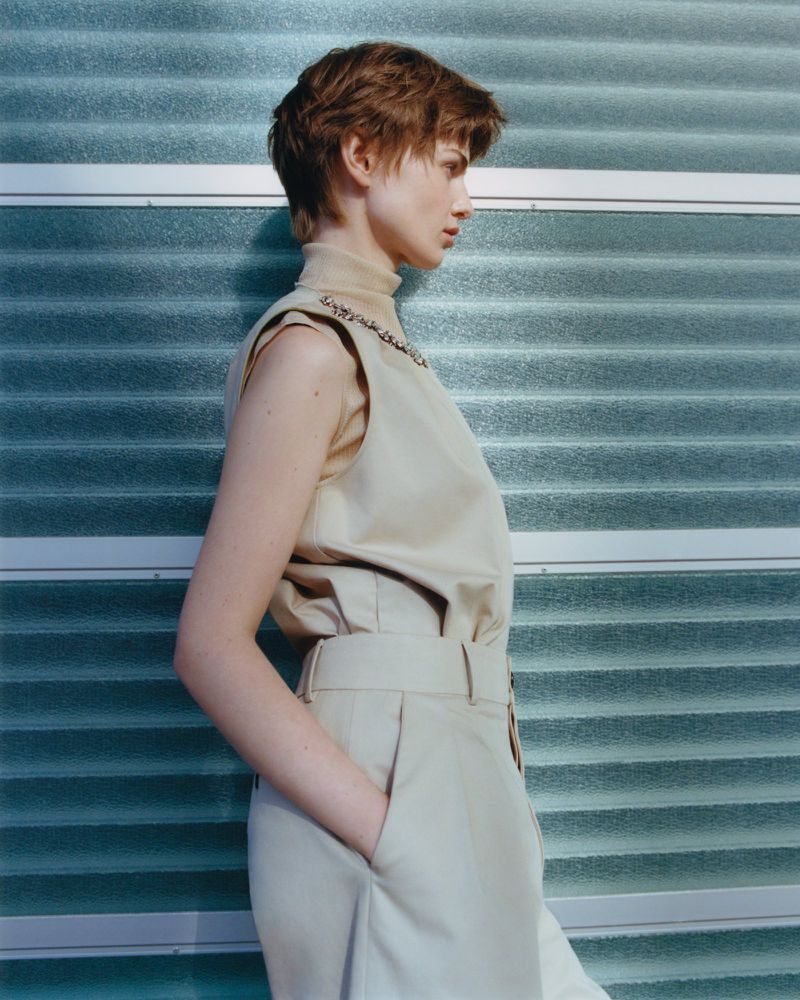 Fashion portrait of a woman with short brown hair in a sleeveless beige top and beige trousers in front of a ribbed blue background.
