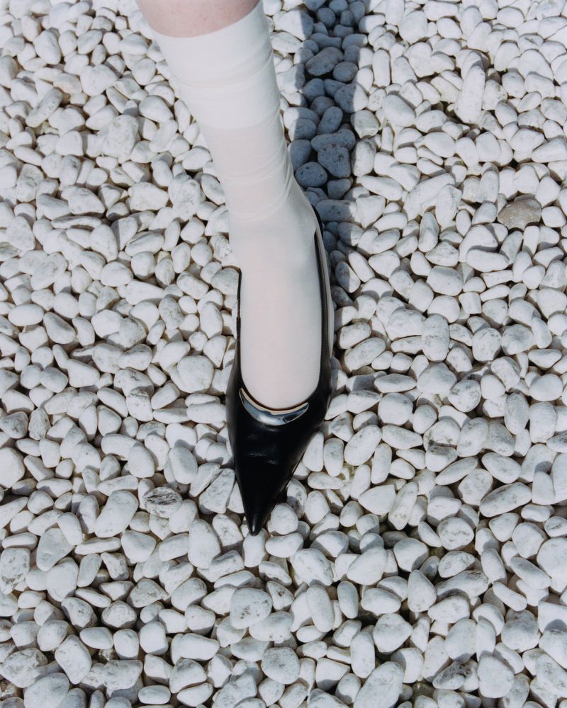 Close-up shot of a person's foot wearing a black pointed-toe high heel and a white sock, standing on small white rocks.