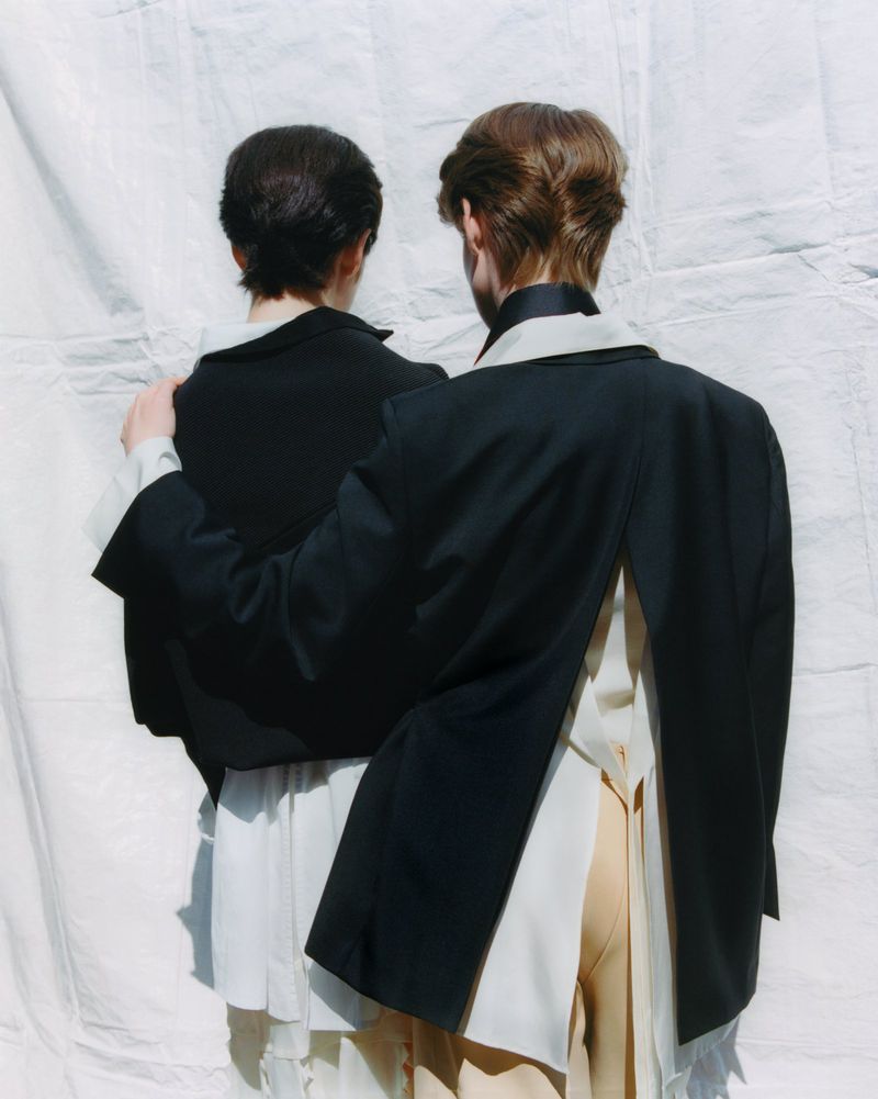 Two people stand close, viewed from behind, wearing black blazers and light-colored shirts and pants, against a white backdrop.