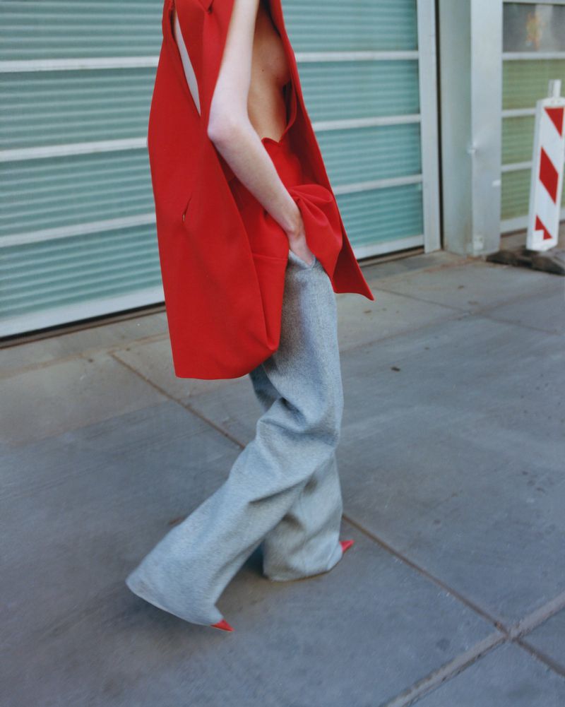 Fashionable woman walking in grey slacks and oversized red vest, with red nails visible, street scene, daylight.