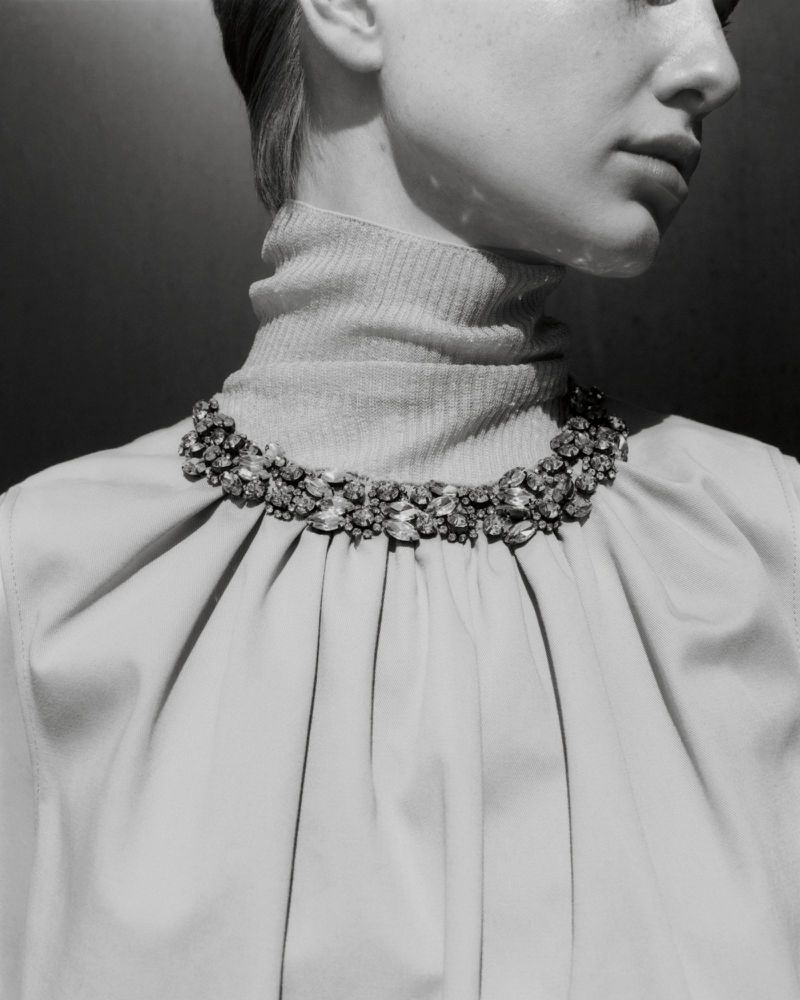 Close-up of an adult female wearing a pale turtleneck and an elaborate jewel necklace, depicted in a black and white studio shot.