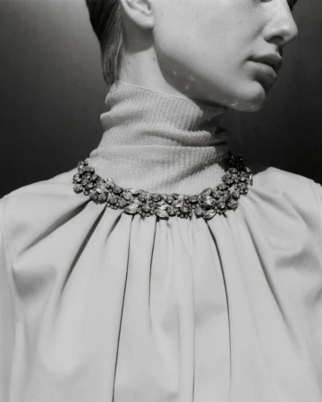 Close-up of an adult female wearing a pale turtleneck and an elaborate jewel necklace, depicted in a black and white studio shot.