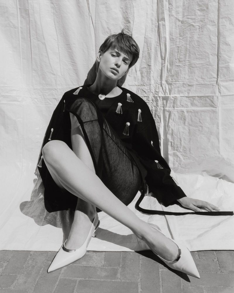 Black and white portrait of a seated, fashionable adult female model wearing a sweater and white heels against a draped white sheet.