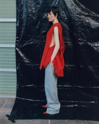 Model with short dark hair wearing a red sleeveless top and grey trousers against a crinkled black plastic sheet background.