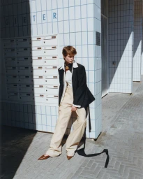 Model wearing a black blazer and wide-leg beige trousers leaning against a white-tiled wall with mailboxes in direct sunlight.