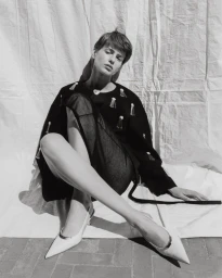 Model with short hair wearing a black embellished top and white pointed-toe kitten heels seated against a textured white backdrop.