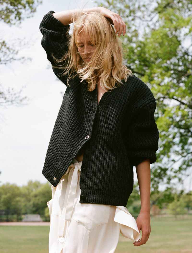 Blonde woman in an oversized black ribbed cardigan and cream skirt touching her hair in an open grassy park.