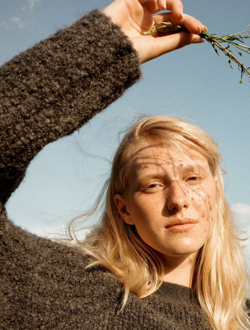 Portrait of a fair-skinned adult woman in a grey sweater, holding green foliage, sky background with shadows cast across her face.