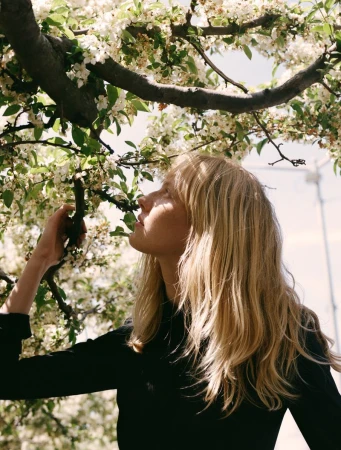 Blonde woman in black long-sleeve shirt standing under a blossoming tree in natural light, profile view, springtime fashion editorial.