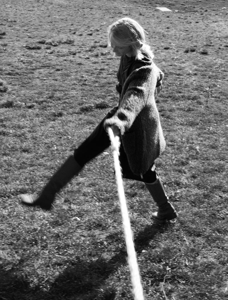 A woman in a coat and boots is outside walking and pulling on a thick rope in a grassy field. The black and white photo has a vintage quality.