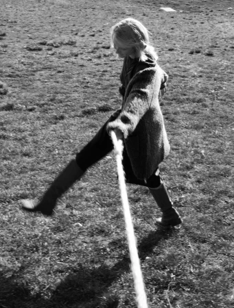 A woman in a coat and boots is outside walking and pulling on a thick rope in a grassy field. The black and white photo has a vintage quality.