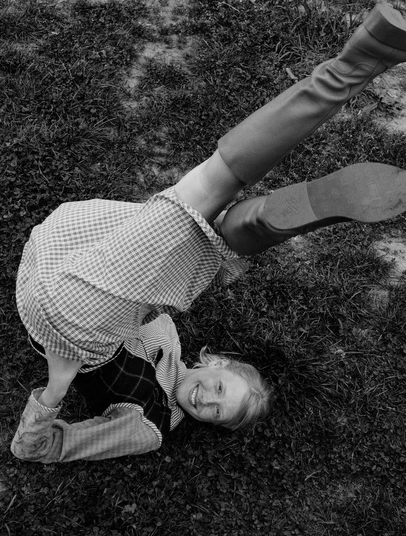 Overhead shot of a young woman lying on grass, legs in the air, wearing boots and a checkered dress, smiling at the camera.