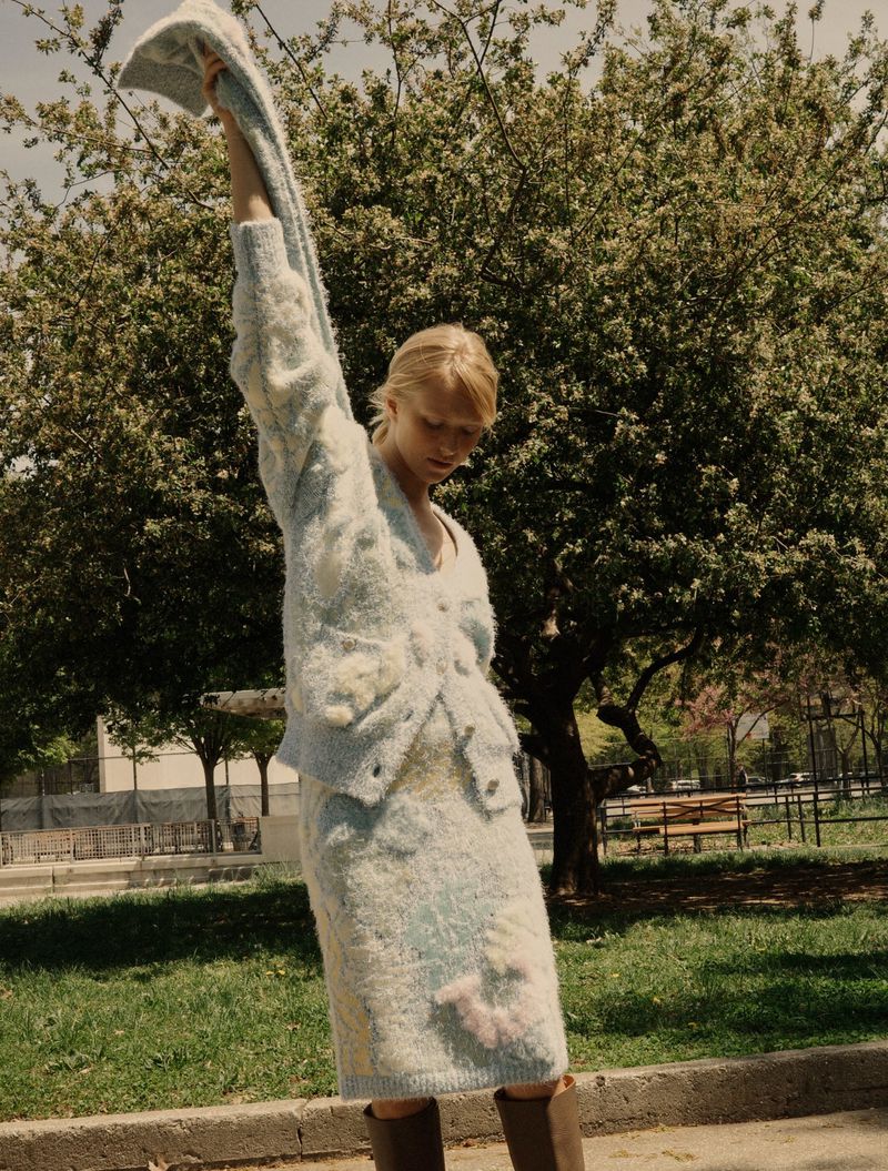 Woman wearing a light blue, floral sweater dress and boots, holding a matching sweater overhead, posing in a park with trees.