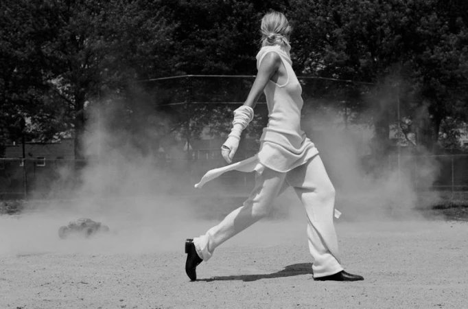 Monochrome full shot of a blonde adult female walking in a field wearing white clothing in front of a cloud of dust on a sunny day.