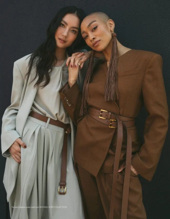 Two fashion models posing closely in tailored grey and brown suits with wide leather belts against a dark seamless backdrop.