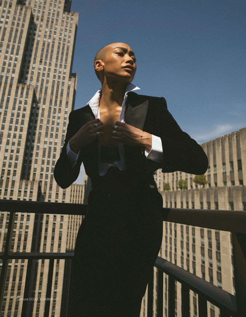 Model with a shaved head in a black tailored suit and open white shirt posing on a balcony before city skyscrapers.