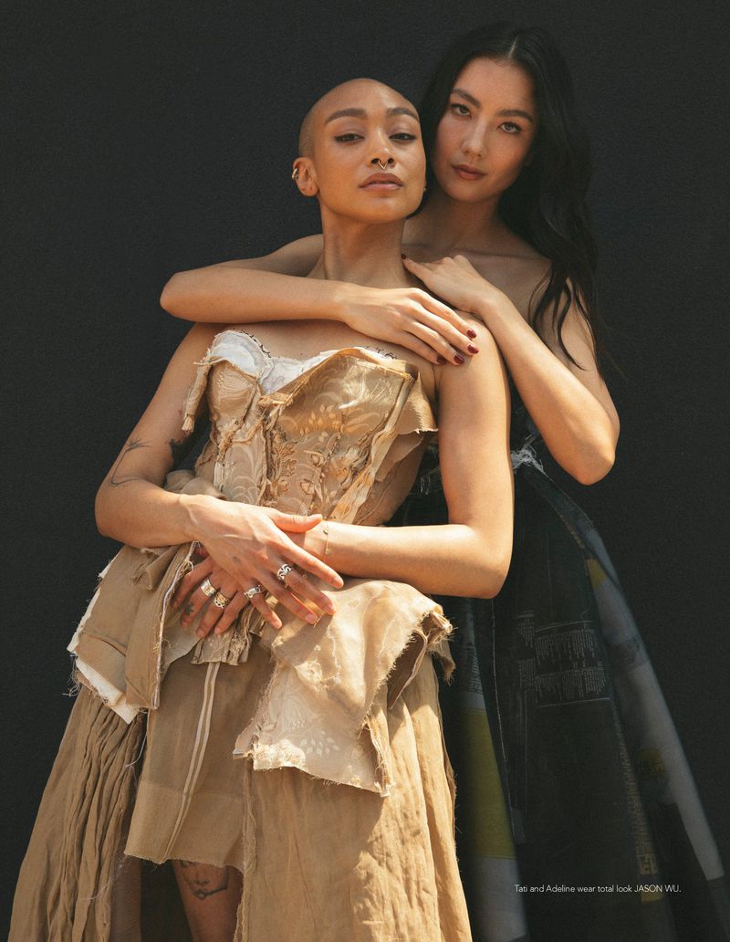 Two women embracing against a black backdrop, one with a shaved head in a beige corset dress and one behind with long dark hair.