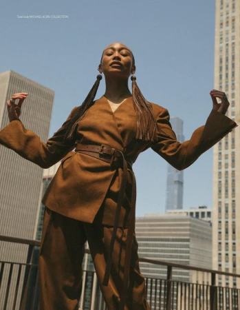 Model in a brown belted suit with long pigtails posing on an urban rooftop against city skyscrapers.