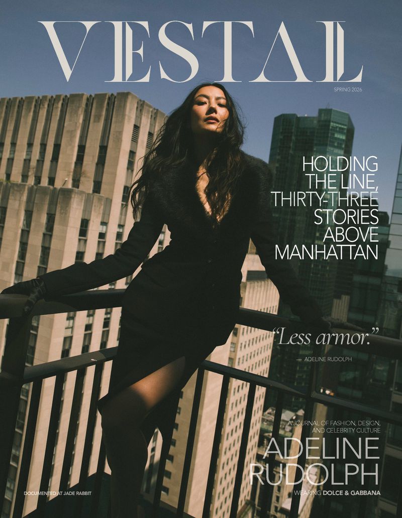 Adeline Rudolph poses in a black coat leaning against a high-rise balcony railing overlooking city skyscrapers on a magazine cover.