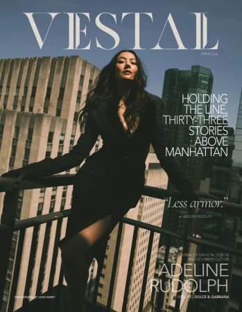 Adeline Rudolph poses in a black coat leaning against a high-rise balcony railing overlooking city skyscrapers on a magazine cover.