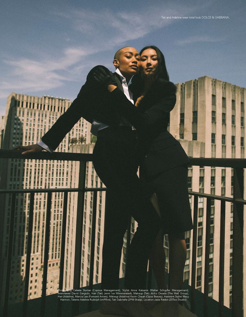 Tati Gabrielle and Adeline Rudolph in black tailored suits posing together on a high-rise balcony overlooking city skyscrapers.