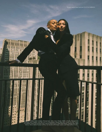 Tati Gabrielle and Adeline Rudolph in black tailored suits posing together on a high-rise balcony overlooking city skyscrapers.