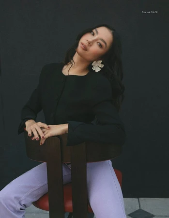 East Asian woman in a black jacket and lilac trousers seated backwards on a wooden chair against a dark backdrop.
