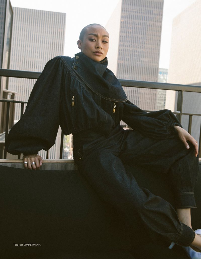 East Asian model with short hair wearing a dark denim outfit with voluminous sleeves on a balcony overlooking city skyscrapers.