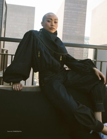 East Asian model with short hair wearing a dark denim outfit with voluminous sleeves on a balcony overlooking city skyscrapers.