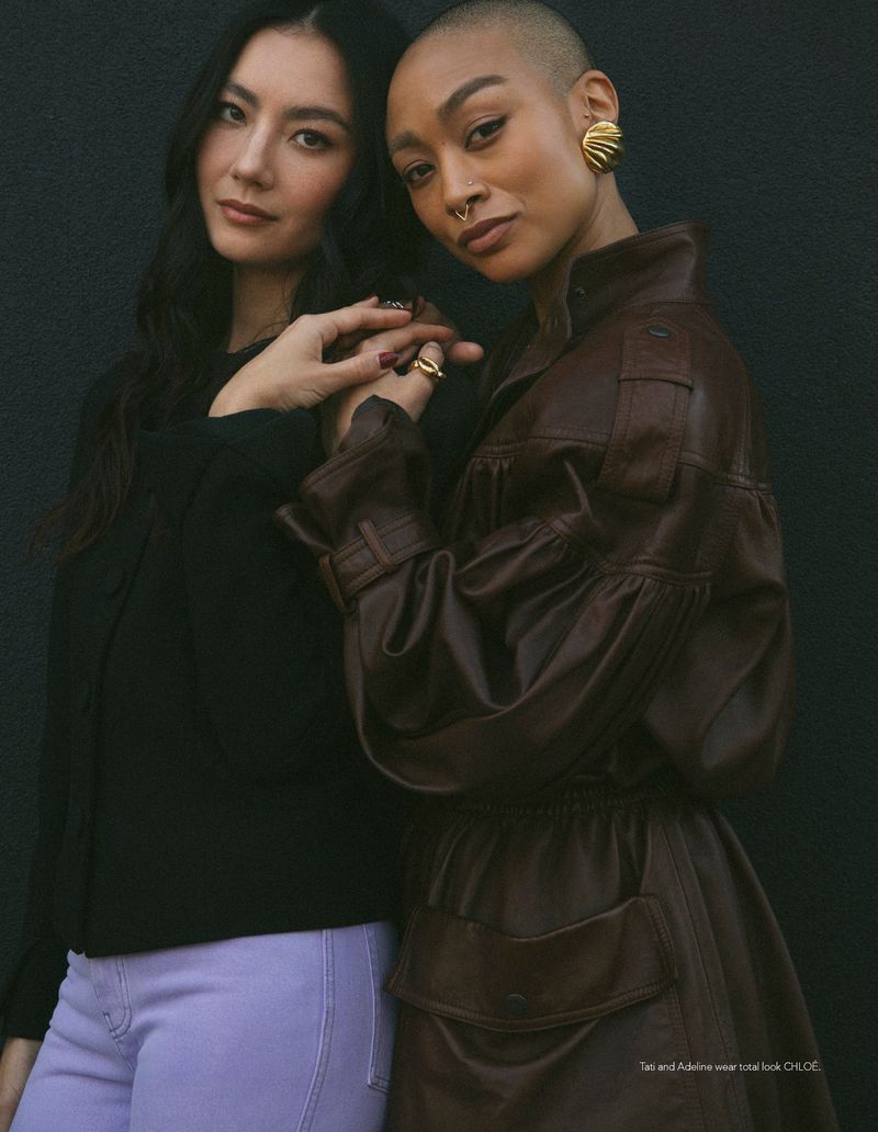 Two women posing closely against a black backdrop, one wearing a brown leather belted coat and gold shell earring.