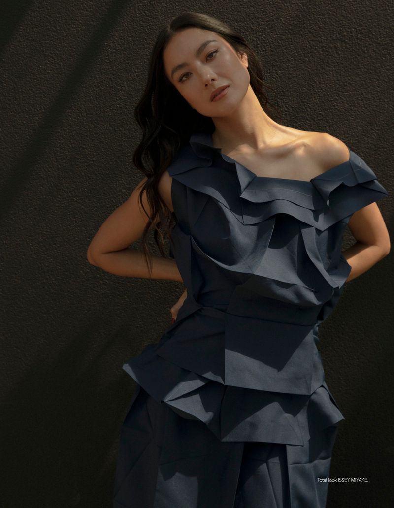 Fashion model wearing a dark sculptural ruffled off-the-shoulder dress against a dark textured background in direct sunlight.