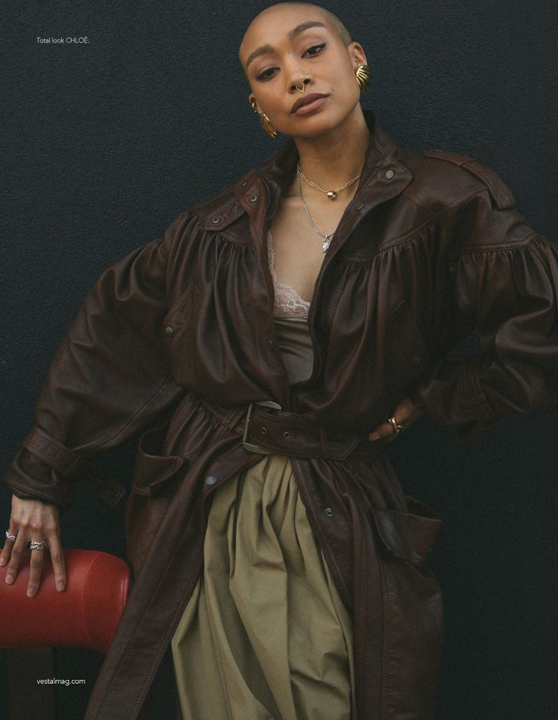 Model with shaved head wearing an oversized dark brown leather coat with gold jewelry against a dark background.