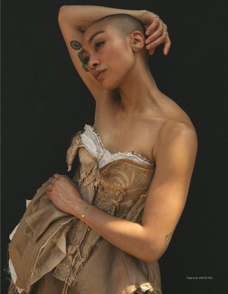 Model with shaved head posing in a deconstructed tan corset dress against a solid dark background.