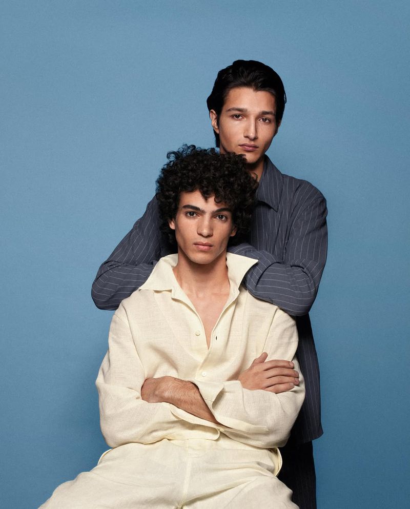 Two male models posing against a solid blue backdrop, one seated in a cream collared shirt and one standing behind in dark stripes.