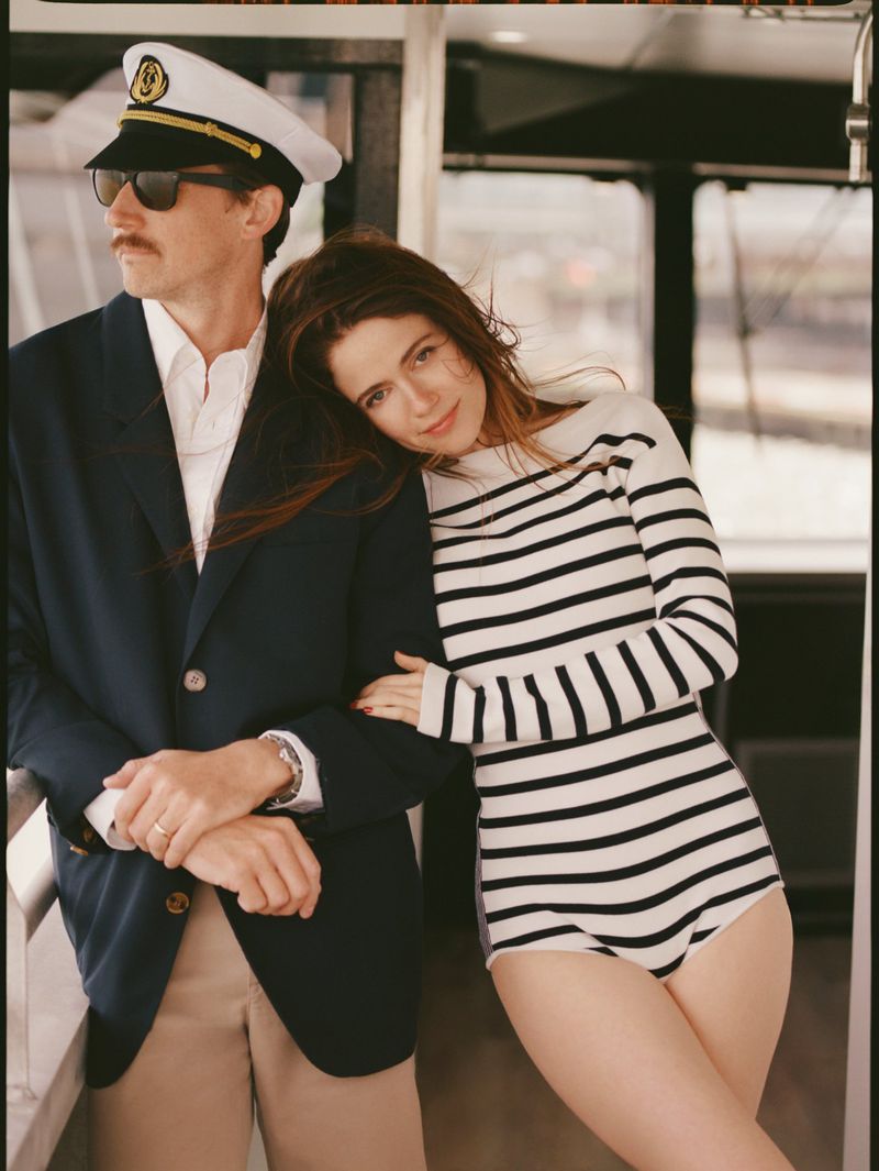 Nautical-themed fashion shoot with a man in a captain's hat and blazer posing with a woman in a striped top and swimwear on a boat.
