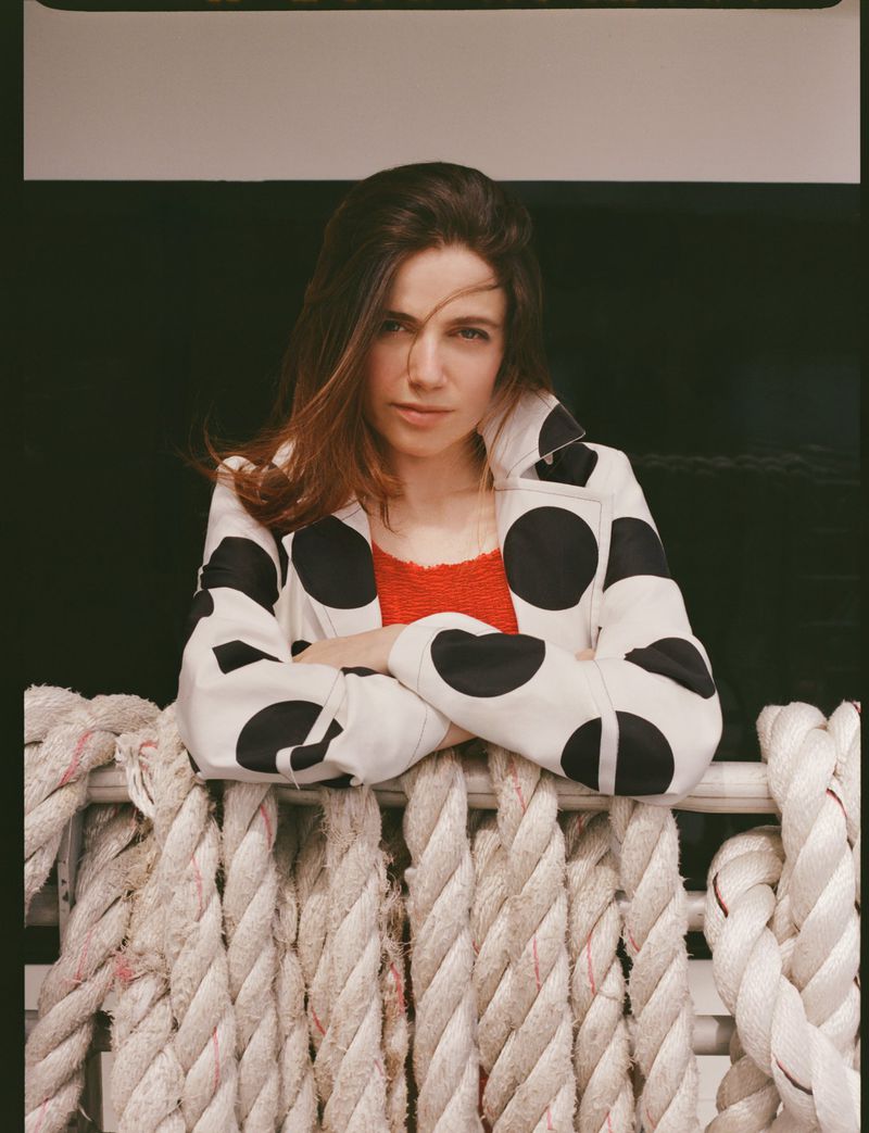 Portrait of a brunette woman wearing a black and white polka dot jacket, leaning on nautical ropes in an indoor setting.