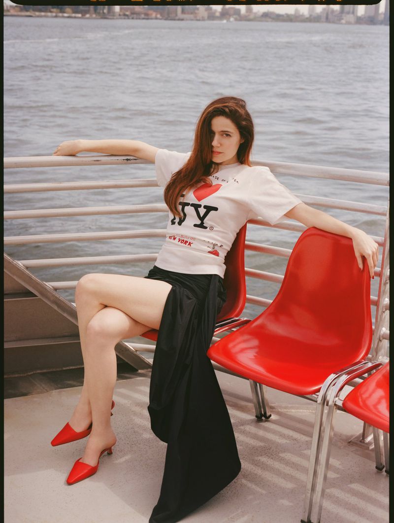 A woman sits on a red chair on a ferry, wearing an I Heart NY shirt, black skirt, and red heels, with city buildings in the background.