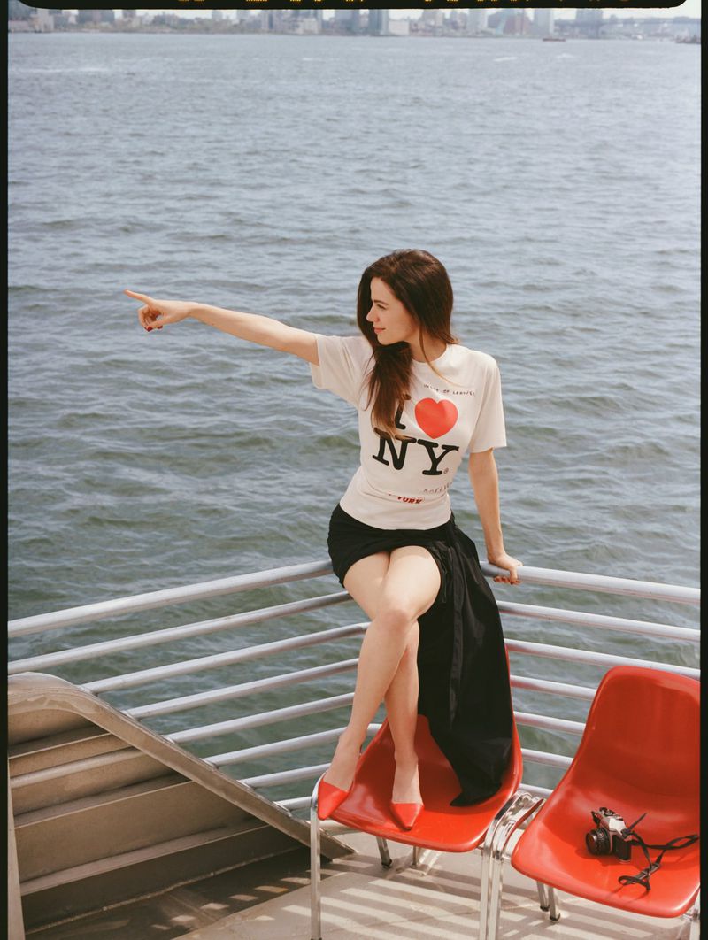 Woman in 'I Love NY' t-shirt sits on a red chair by the water, pointing. River, buildings, and city skyline in background on a sunny day.